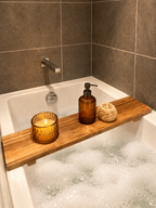 A wooden bath caddy draped over a bubbling bathtub, holding an amber glass candle holder, an amber pump bottle, and a natural loofah scrubber, set against dark gray tiled walls.