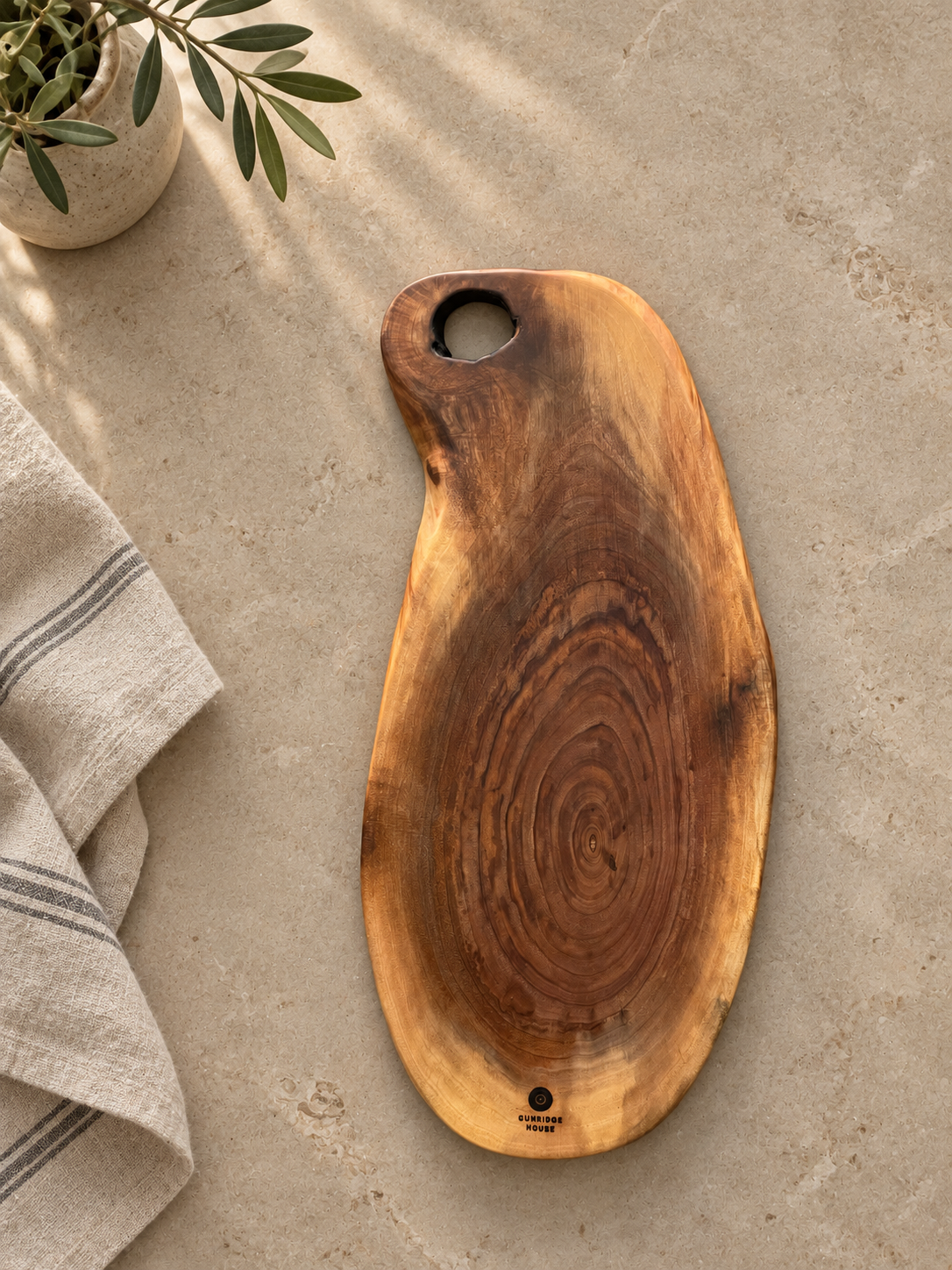 A rustic live-edge wooden serving board with a natural grain pattern and a circular cutout at the top, styled on a textured beige stone surface with a striped linen towel and a small potted plant.