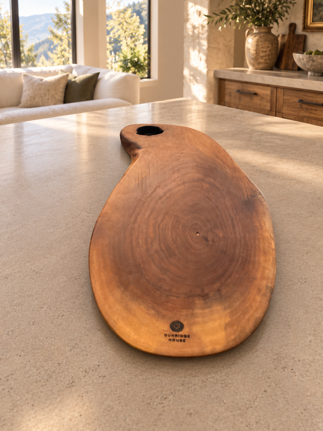 A large organic-shaped walnut wood serving board with a natural live edge and a circular cutout near one end, placed on a light stone kitchen island. The board has visible wood grain and a small engraved brand mark near the bottom.